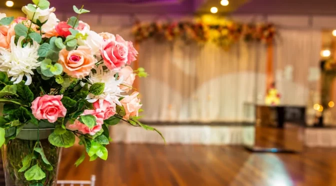 Elegant wedding floral centrepiece with pink, peach and white roses arranged in a glass vase at a reception venue.